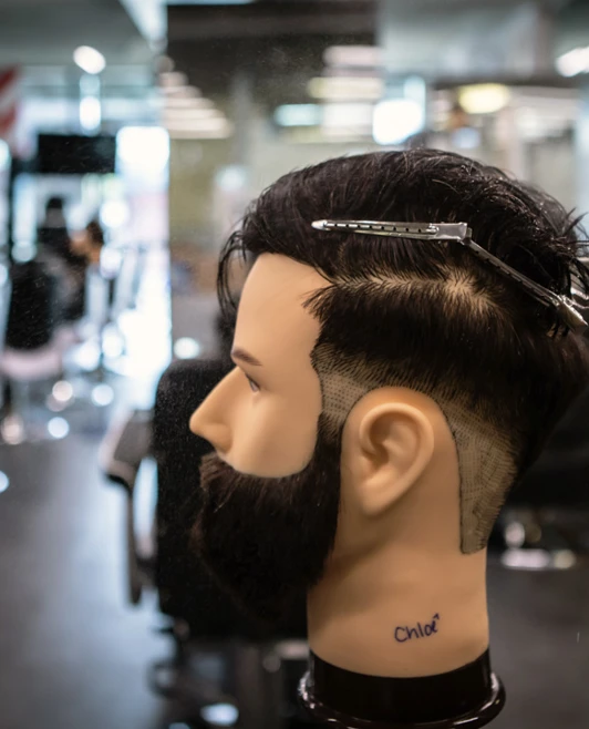 A mannequin head with a beard and a partially completed haircut in a barbering academy, with a hand spraying water onto the hair. A mannequin head with a beard and a partially completed haircut in a barbering academy, with a hand spraying water onto the hair.